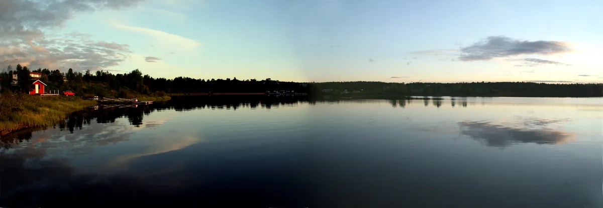 Calm, mirror-still waters of Lake Inari reflecting the sky at midnight sun, with a red cabin and wooded shoreline, Finnish Lapland