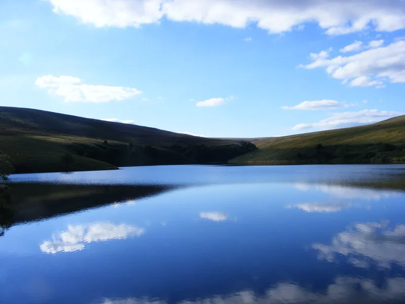 Calm reflective water of Grwyne Fawr Reservoir surrounded by grassy Black Mountains hillsides under a partly cloudy sky.