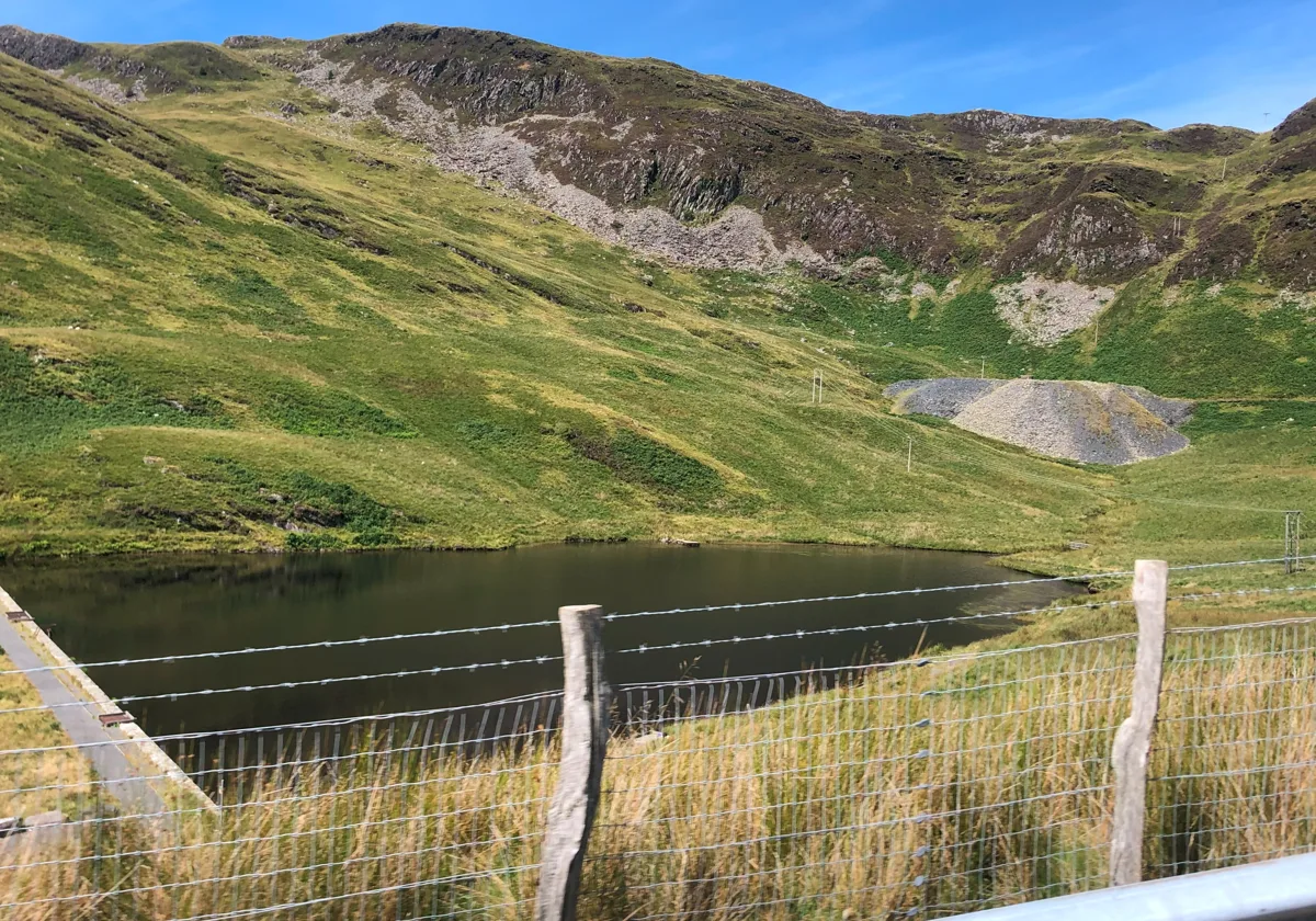 Small upland reservoir lake (Llyn Ffridd-y-Bwlch) in Snowdonia surrounded by green hillside, with a wire fence in the foreground.
