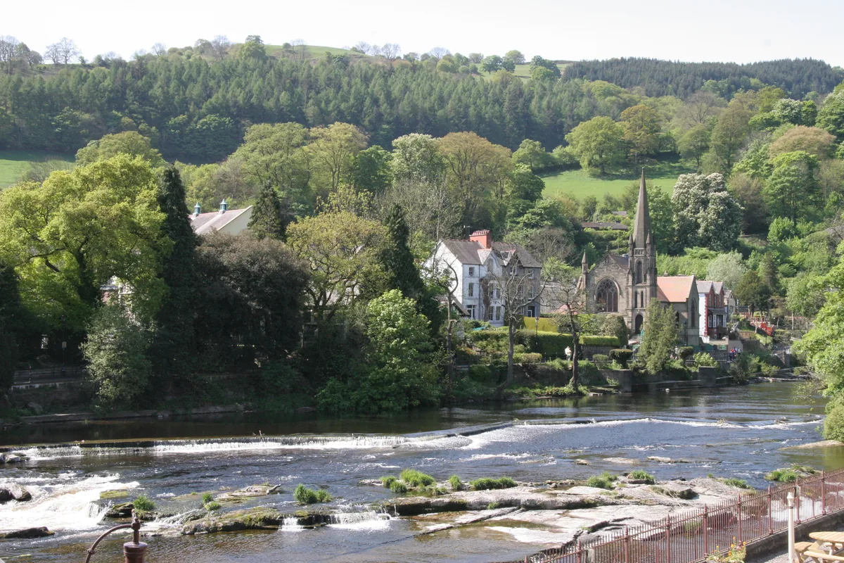 Wide view of the River Dee flowing over a weir through the town of Llangollen, Denbighshire, Wales, with wooded hills behind