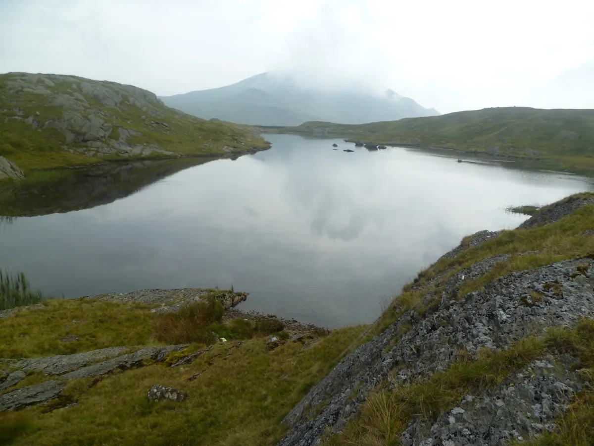 Calm upland lake (Llyn Cwm Corsiog) surrounded by rocky, grassy banks with misty hills in the background, Snowdonia.