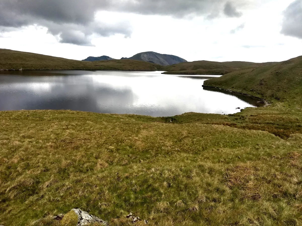 Still upland lake (Llyn Conglog) reflecting a cloudy sky, with grassy moorland banks and a mountain in the distance, Snowdonia.