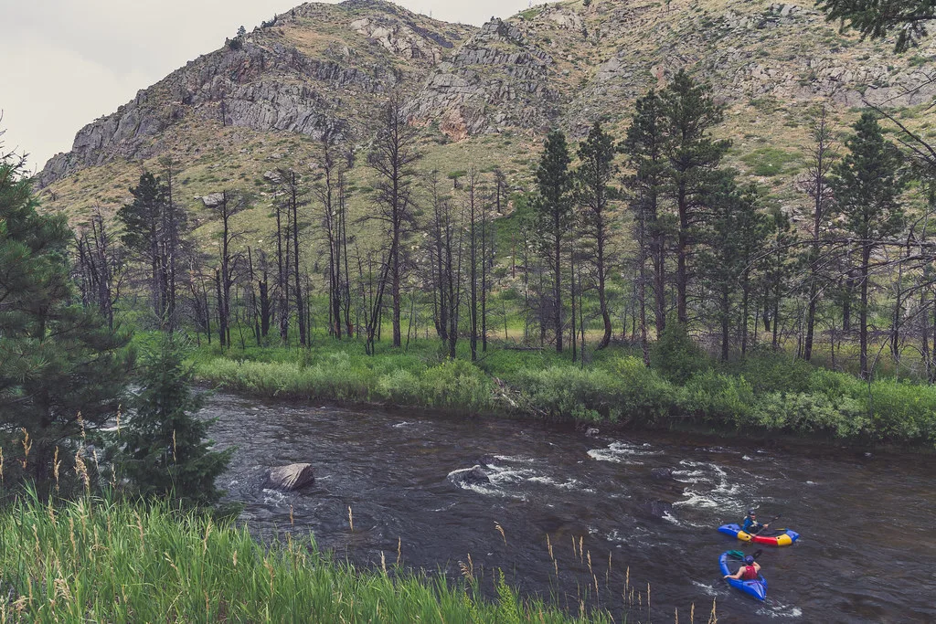 The Cache la Poudre River flowing through a rocky canyon lined with pines below the Colorado Front Range, with two rafters on the water.