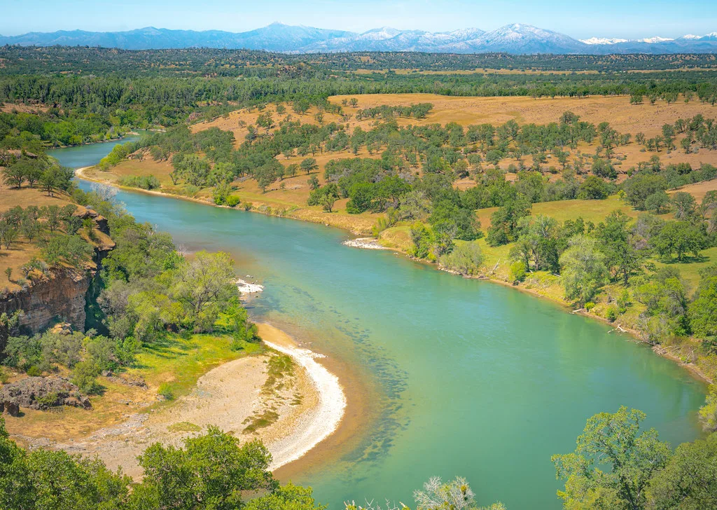 Aerial view of the turquoise Sacramento River bending through oak grassland with mountains in the distance.