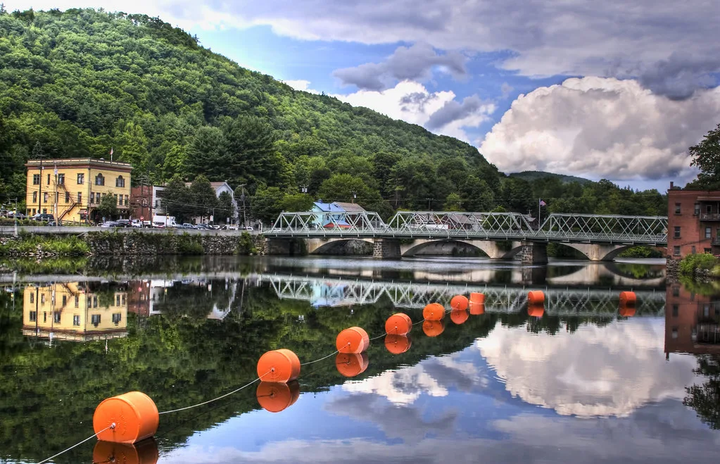 Still water on the Deerfield River reflecting a bridge, riverside buildings, and wooded hills in Massachusetts.