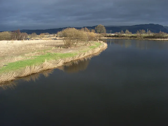 Wide view of the River Forth flowing past grassy riverbanks near Bridge of Allan, with hills in the distance under a dramatic sky.