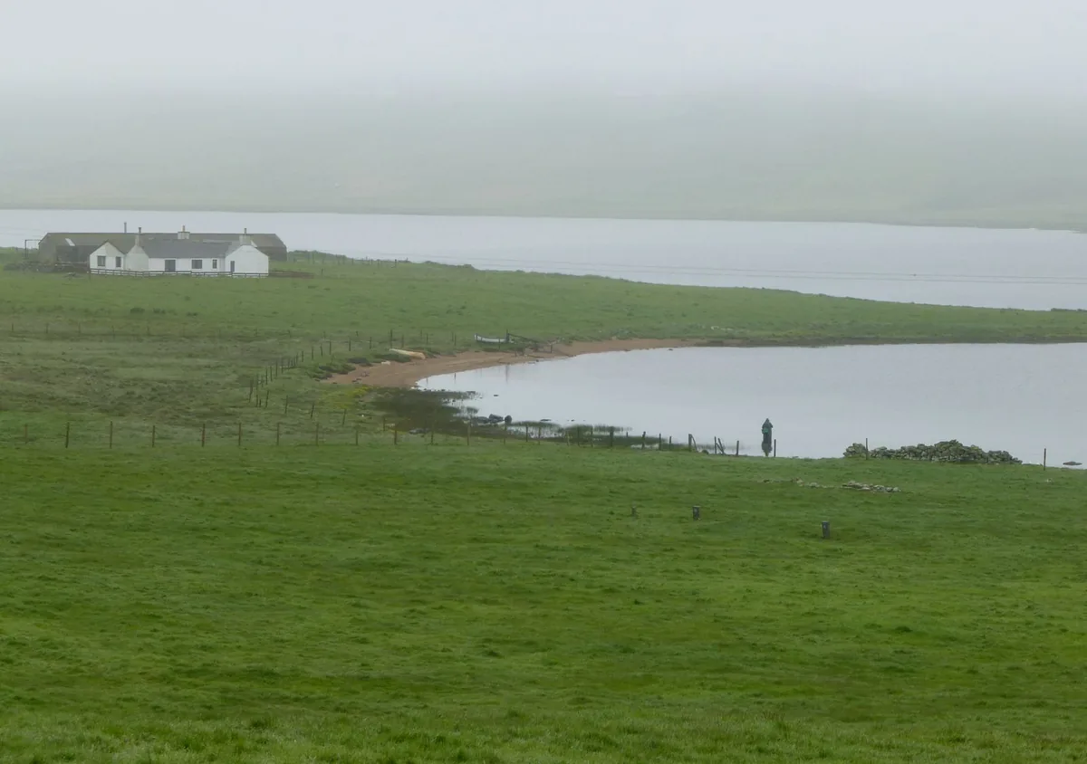A lone fisherman casts from the shore of the Loch of Spiggie in Shetland, with open water and low hills in the background.