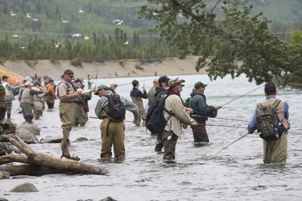 A row of fly anglers wading and casting at the confluence of the Kenai and Russian Rivers, with gulls flying overhead and forested banks behind.