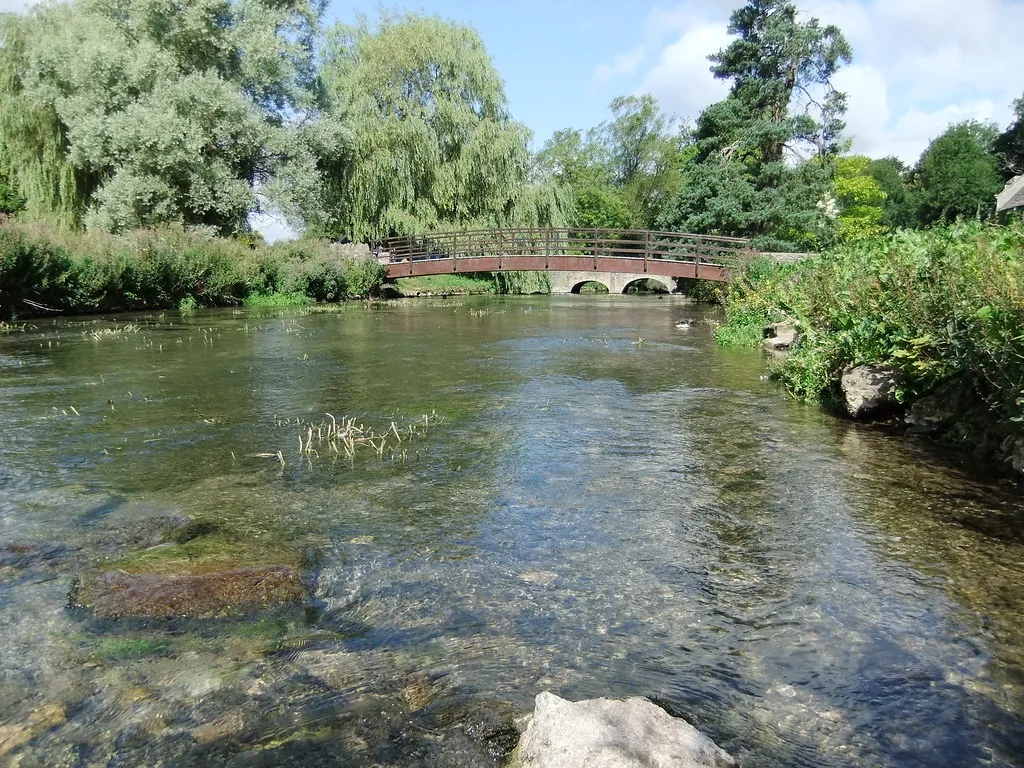 A clear chalk stream, the River Coln, with visible gravel bed and weed, flowing under an arched footbridge lined with willows.