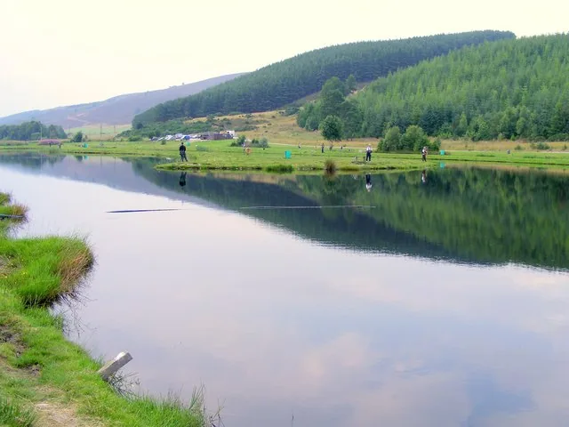 Wide view of a calm fishing pool at Glen of Rothes trout fishery, with anglers on the far bank and wooded hills beyond