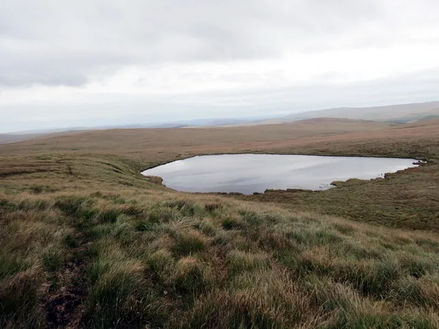 A dark oval upland lake set in open grass moorland under a partly cloudy sky, with distant hills on the horizon.