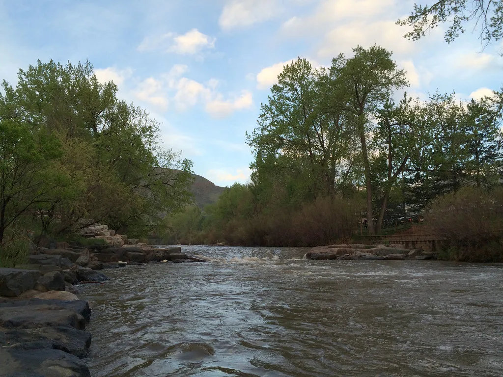 Clear Creek flowing over a small rapid lined with boulders in Golden, Colorado, foothills visible in the background.