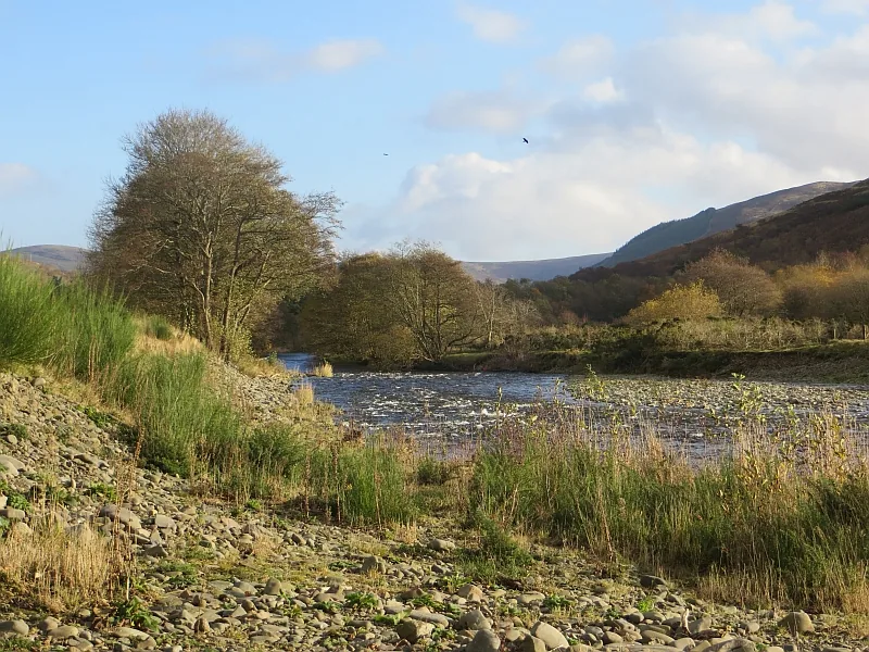 A wide, shallow stretch of Yarrow Water flowing over gravel with a stony bank in the foreground and hills rising beyond.