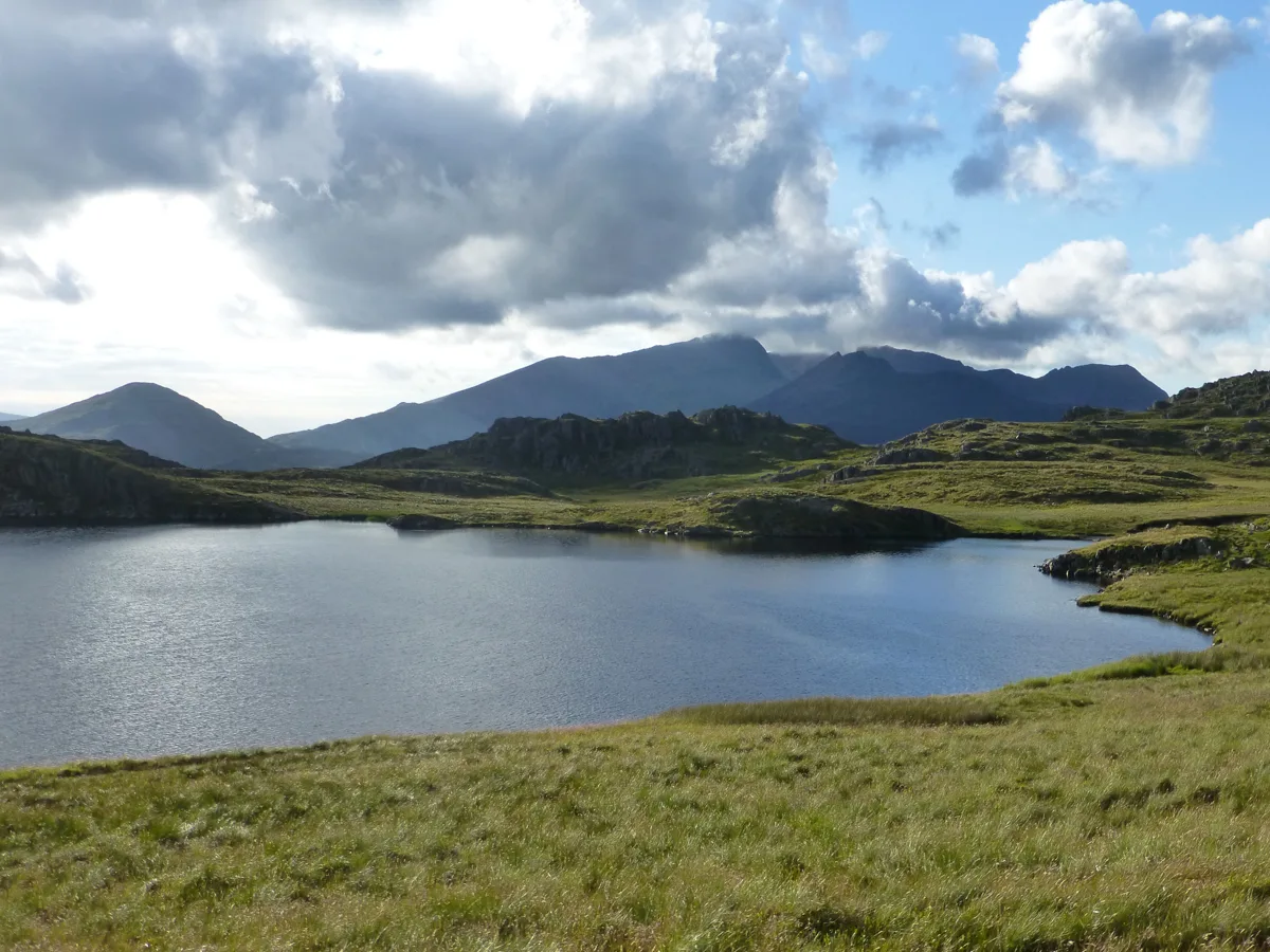 A calm upland lake surrounded by grassy moorland with rocky outcrops, with the mountains of Snowdonia including Snowdon visible under a partly cloudy sky.