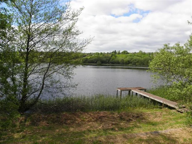 View across the calm water of Corranny Lough towards a wooden fishing jetty and wooded far shore