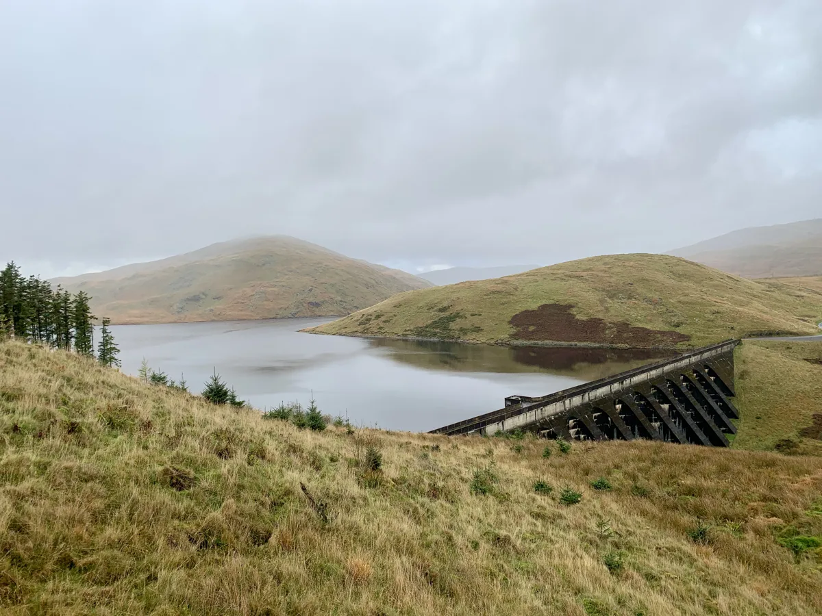 Misty view over Nant y Moch Reservoir in mid Wales, with the concrete dam structure on the right and rolling Cambrian Mountains hills surrounding the water.