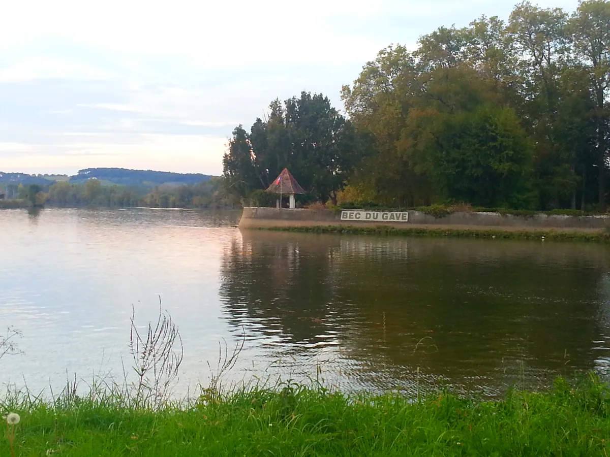A wide, calm river at the confluence of the Adour and the Gaves Réunis, with trees and a small riverside gazebo marked Bec du Gave in the distance under an evening sky.