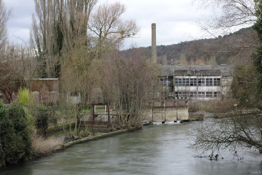 A clear, gently flowing river passing an old stone mill building in a wooded Normandy valley.