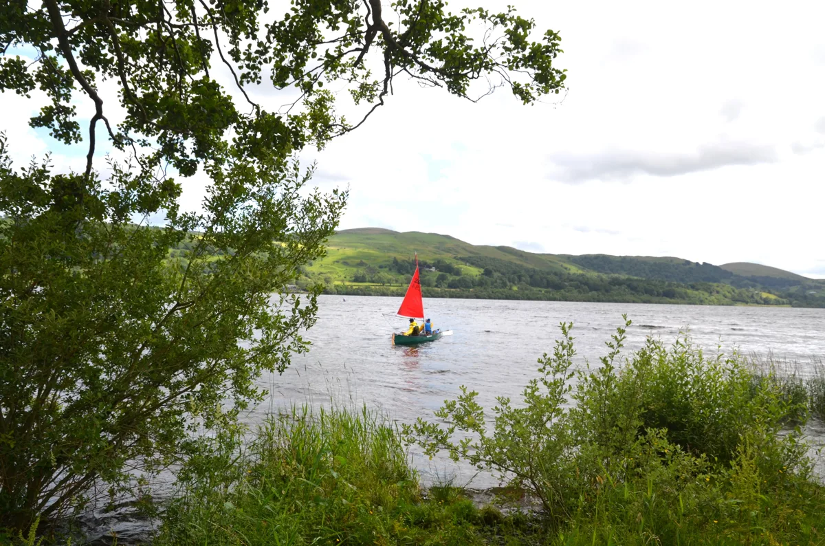Wide view of Bala Lake (Llyn Tegid) from the Llanycil shoreline, with a small sailing boat on the water and hills in the background, framed by lakeside trees.