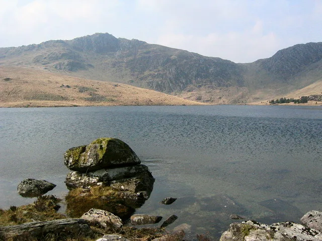 A moss-covered rock in the foreground of a calm upland lake, with bare mountain slopes rising behind under a clear sky.