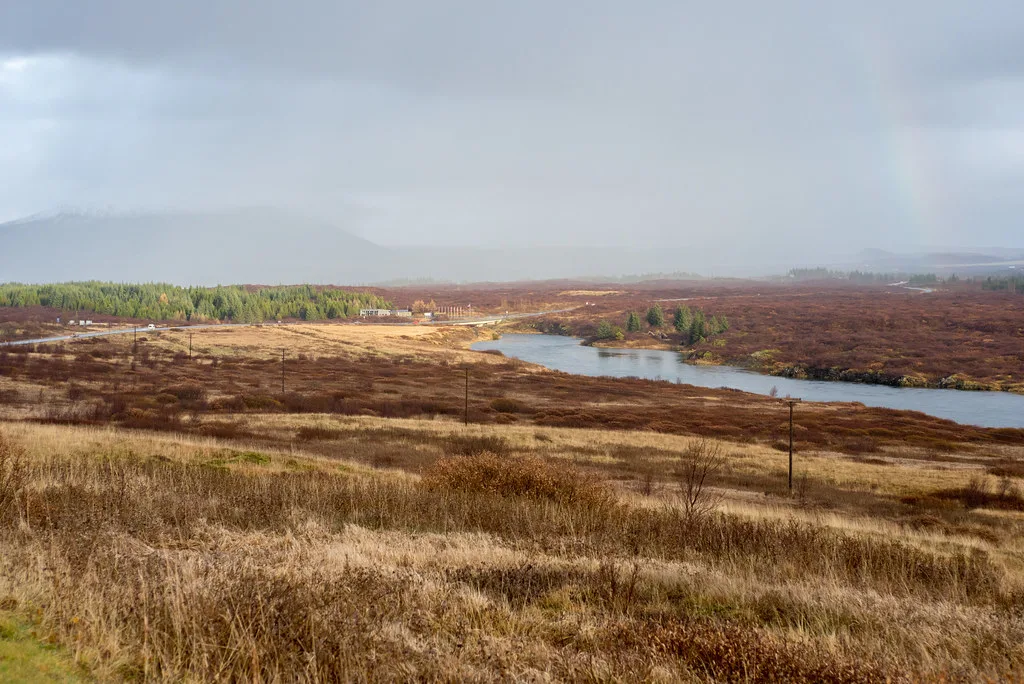 A wide river bending through open moorland with a distant road bridge and mountains, autumn colours in the vegetation, Sog river Iceland.