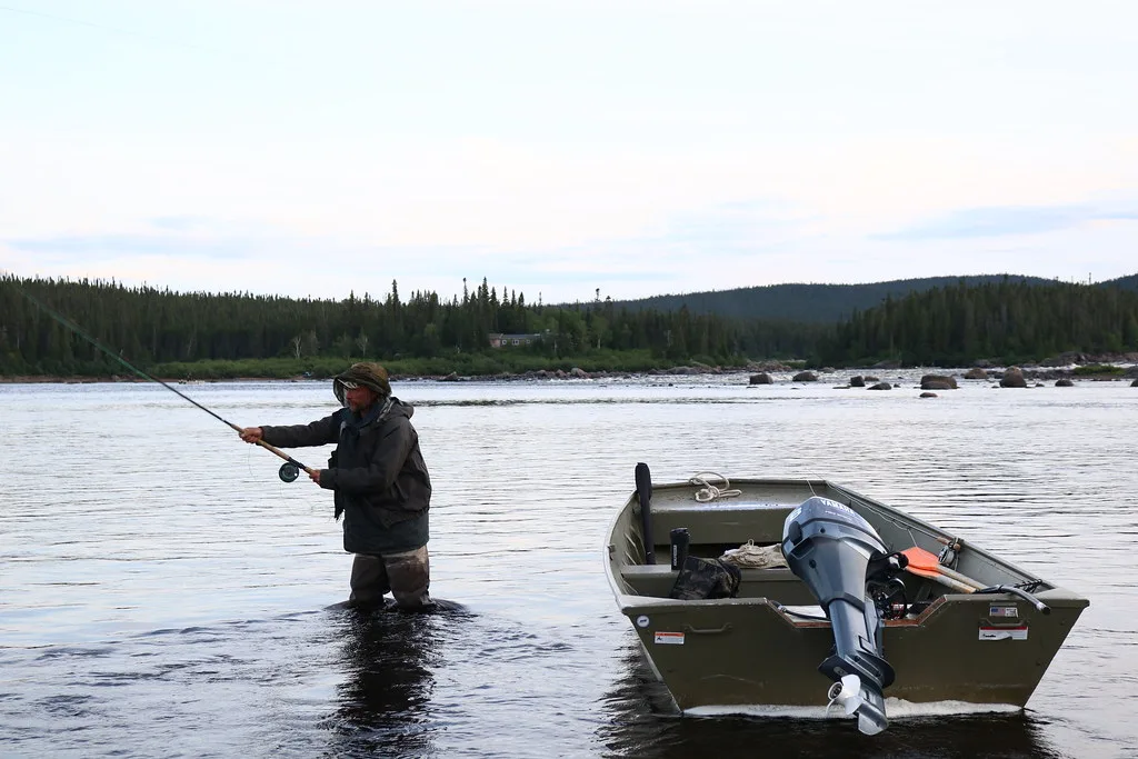 An angler wades the wide Eagle River in Labrador, casting a fly rod beside a jon boat, with boreal forest along the far bank.