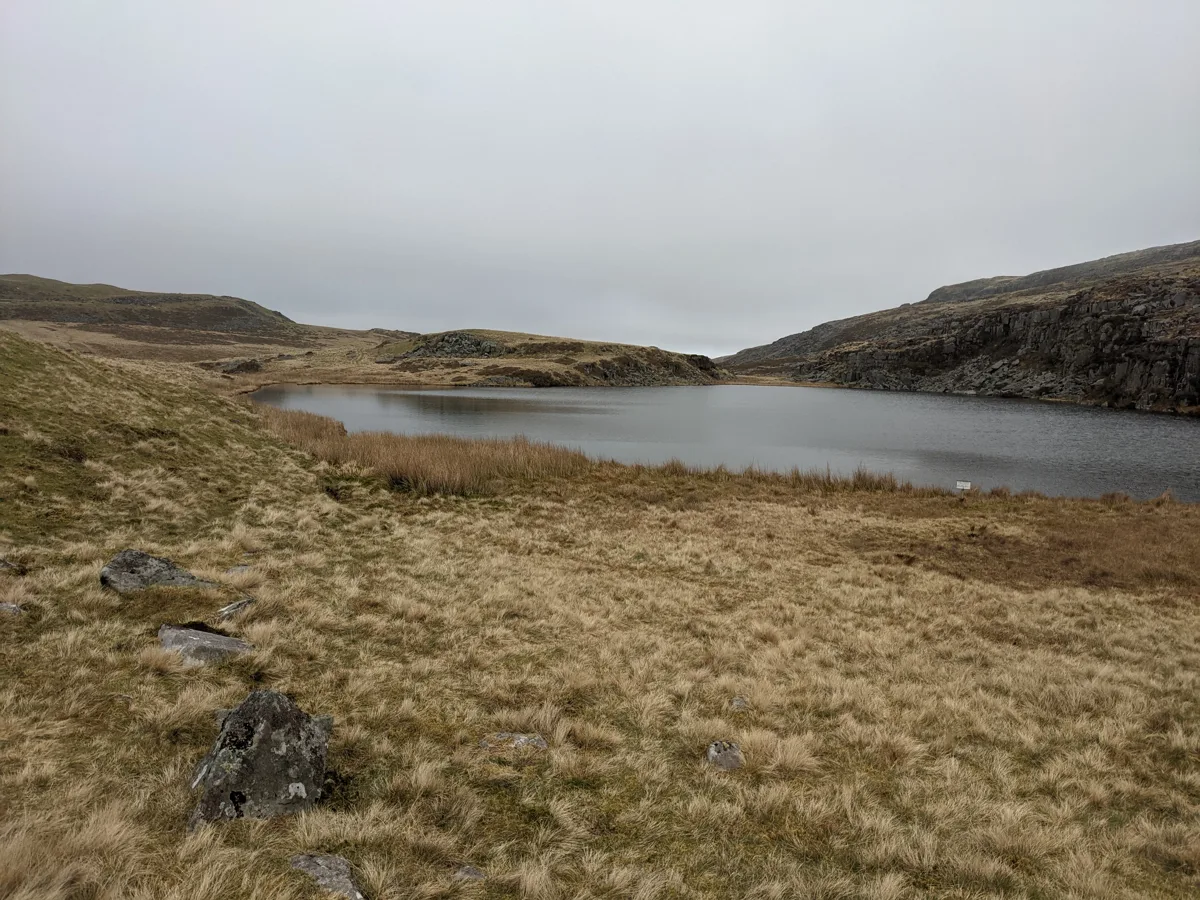 A grey, still upland lake bordered by tussocky moorland grass and a low rocky outcrop, under an overcast sky.