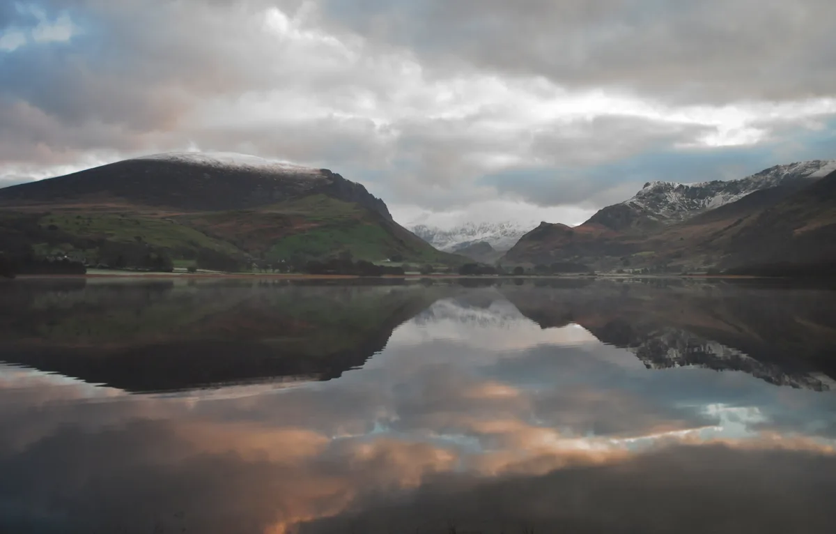 Calm mirror-still Llyn Nantlle reflecting snow-capped Snowdonia mountains and a cloudy pink-tinged sunrise sky.