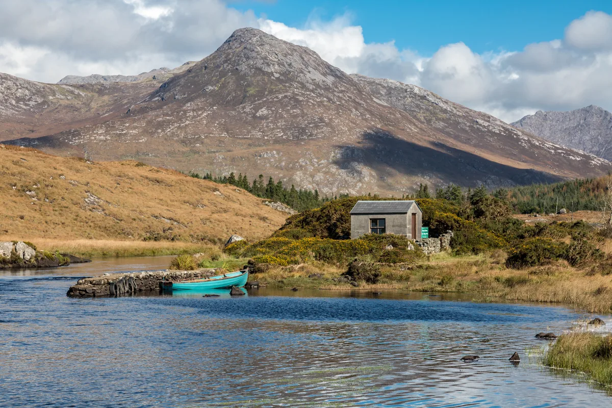 A small stone fishing hut and moored boat beside a lake on the Ballynahinch Fishery estate, Connemara, with the mountain Benlettery in the background