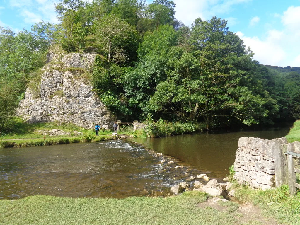 Shallow limestone river with stepping stones and a dry-stone wall, backed by a wooded limestone crag at Dovedale, Derbyshire.