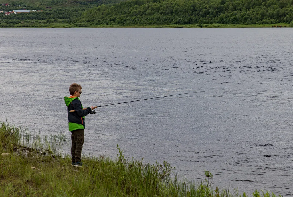 A boy fishing from the grassy bank of a wide river with forested far shore.