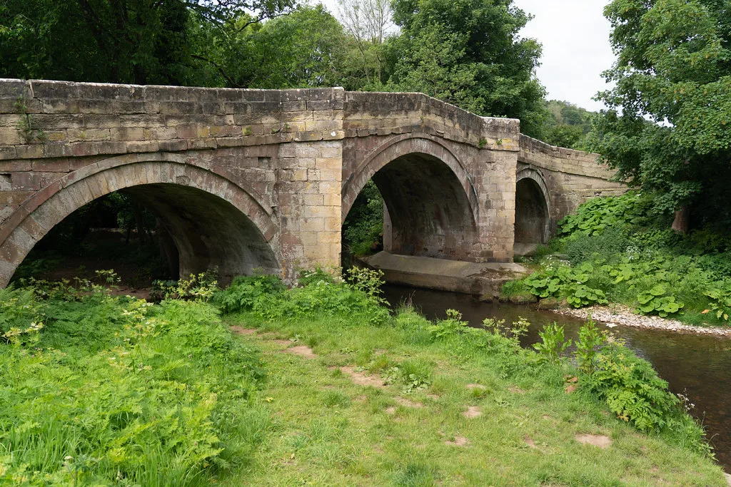 A three-arched stone bridge over the River Rye, surrounded by lush green riverside vegetation near Rievaulx, North Yorkshire.