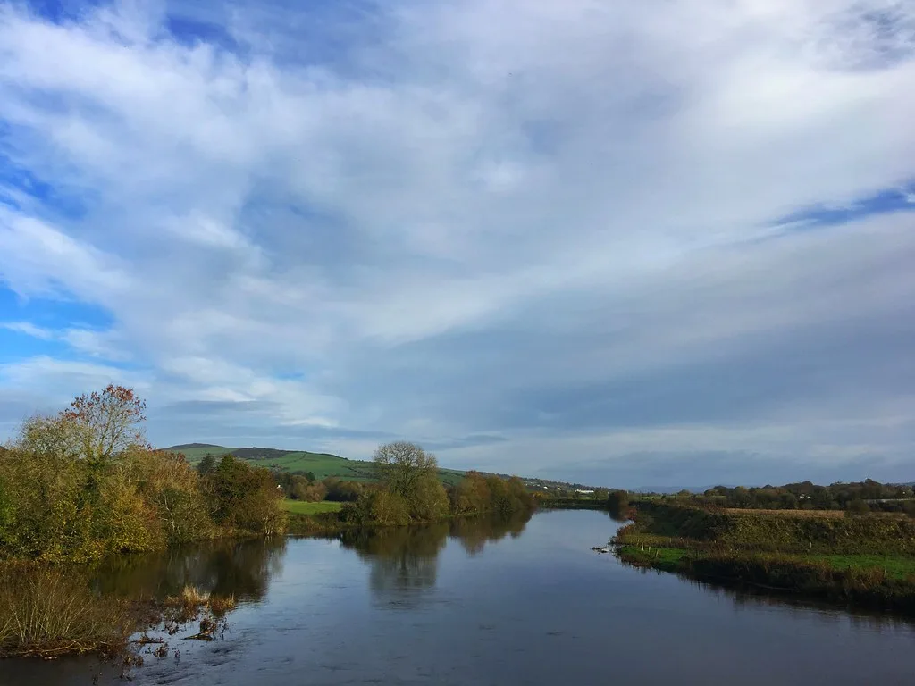 A wide, calm stretch of the River Finn near Clady bridge, flanked by autumn trees and low hills under a cloudy sky.