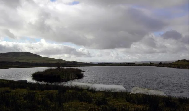 Wide view of the Glenquicken Fishery trout loch under a cloudy sky, with a small island and moorland hills beyond