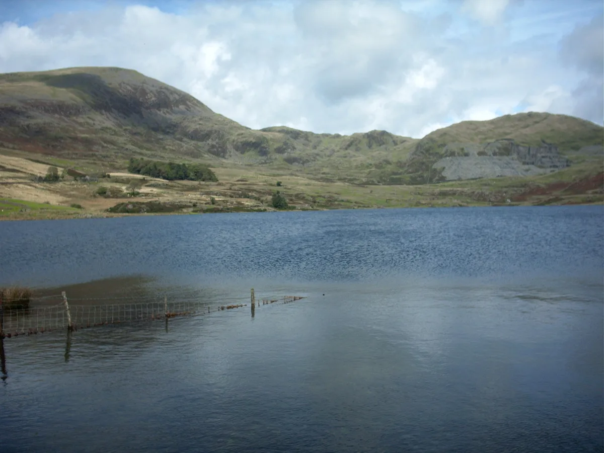 Still blue water of Llyn Cwmystradllyn with a submerged boundary fence, green hills, and an old slate quarry scar on the far hillside under a cloudy sky.