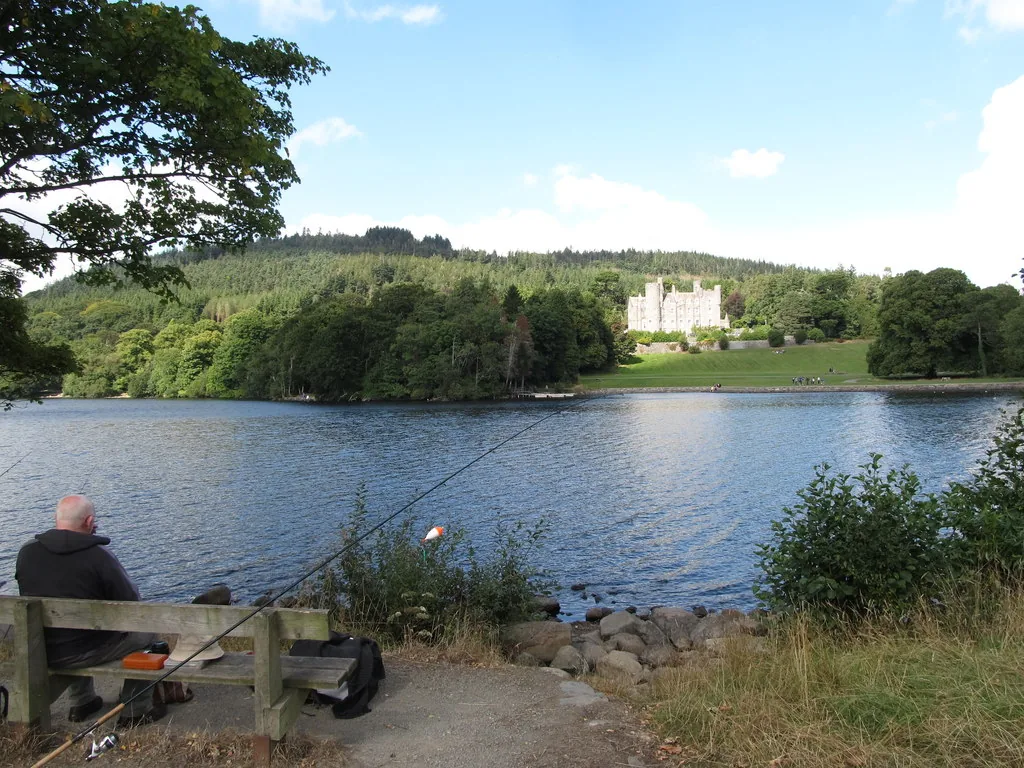 A fisherman seated on a bench fishing the shore of Castlewellan Lake, Castlewellan Castle visible across the water