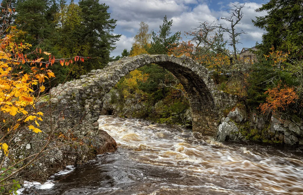 The River Dulnain flowing fast and peat-stained under an old stone packhorse bridge at Carrbridge, surrounded by autumn trees.