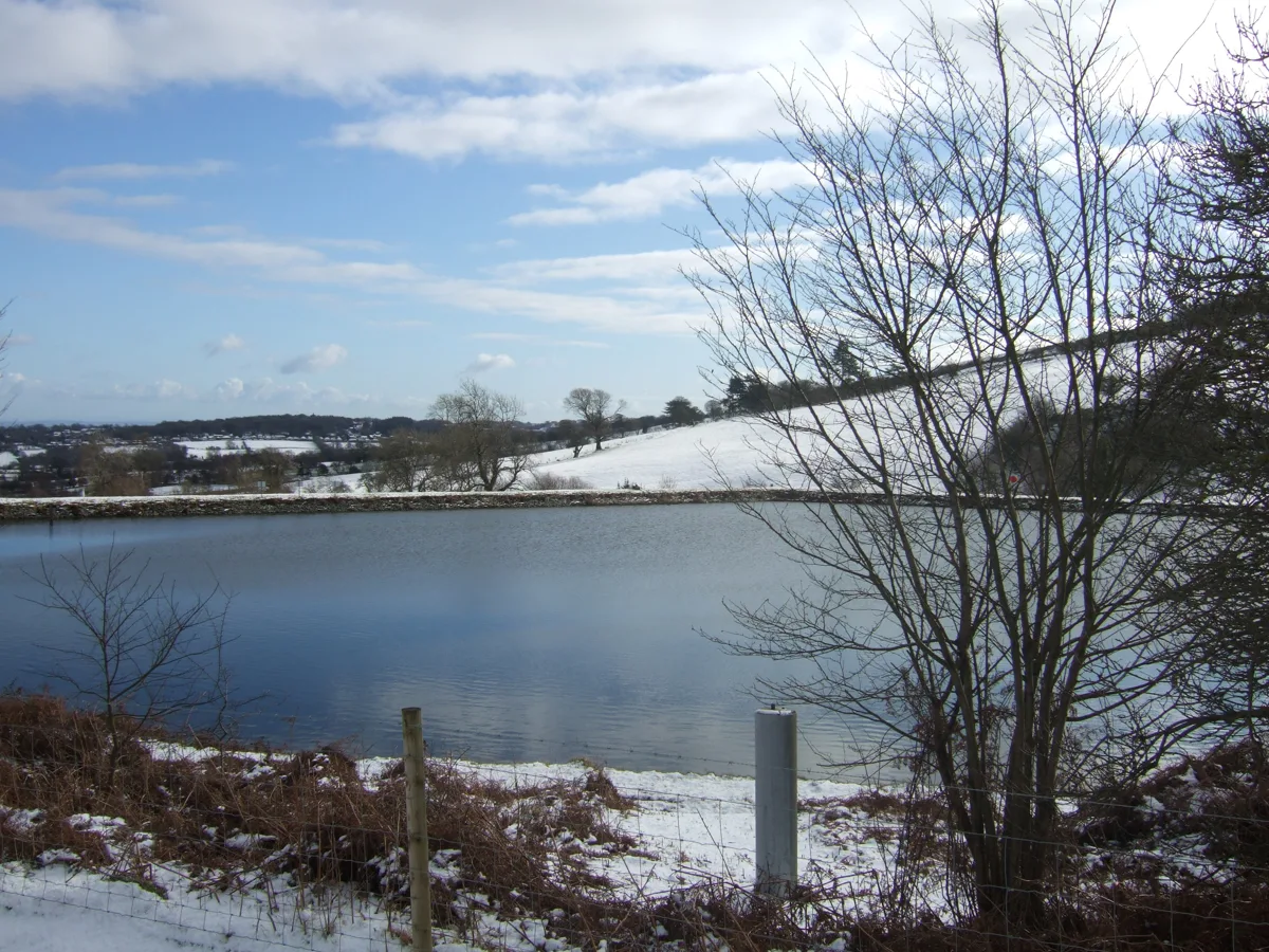 A small reservoir lake with an embankment dam, surrounded by snow-covered fields and bare trees, near Cilcain in the Clwydian Range.
