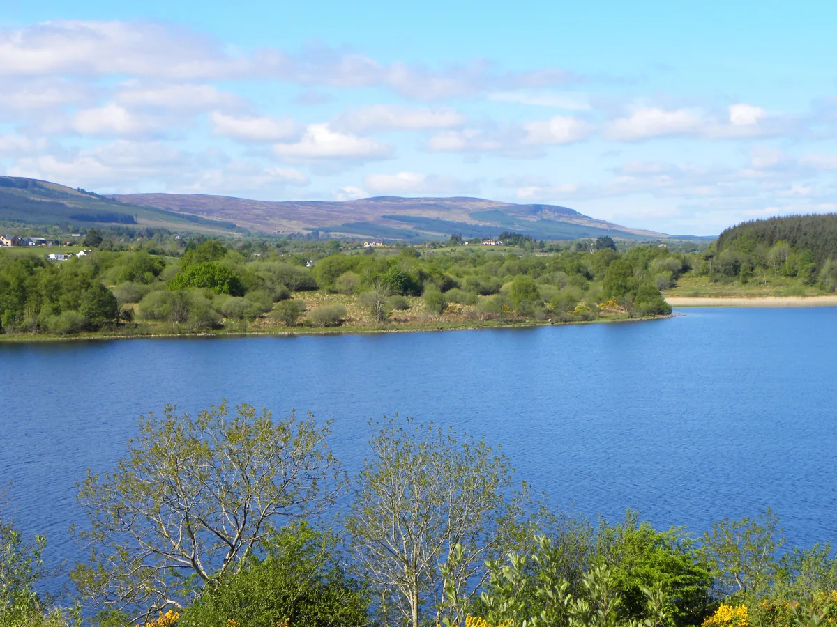 Wide view across the blue water of Upper Lough MacNean towards wooded shoreline and distant hills under a partly cloudy sky.