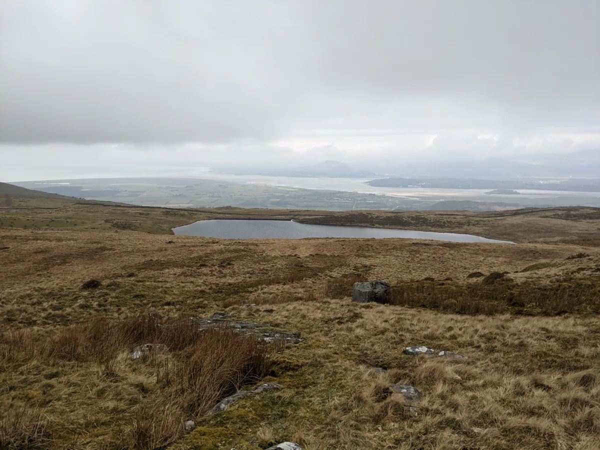 A small elongated moorland lake seen from a heather-covered hillside, with a flat coastal plain and misty hills in the far distance.