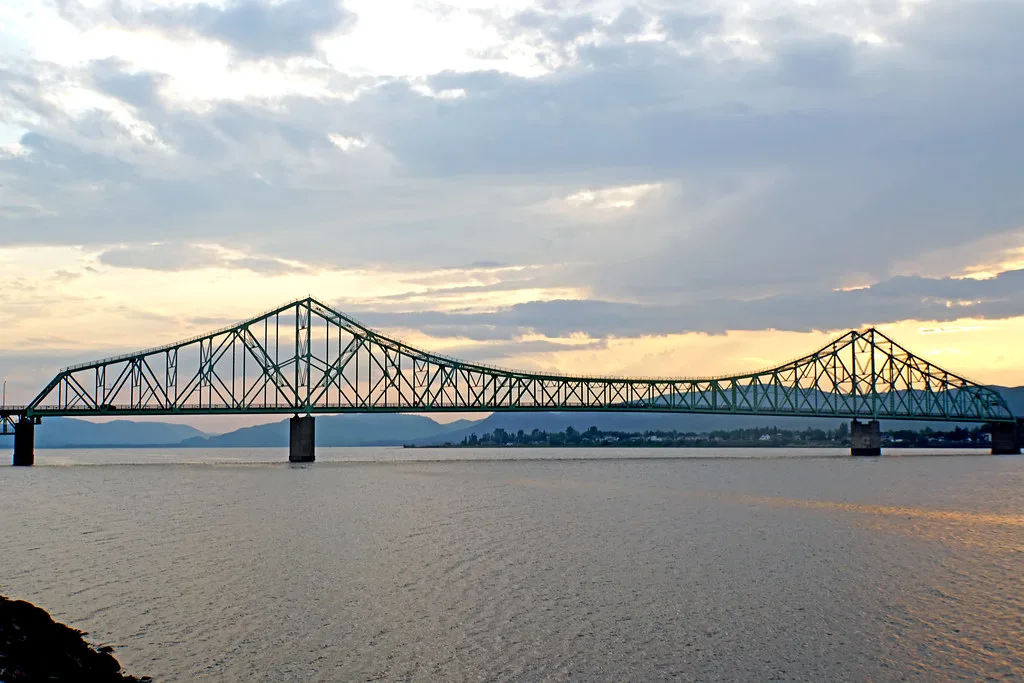 A large steel truss bridge spanning the wide Restigouche River at sunset, with mountains in the background.