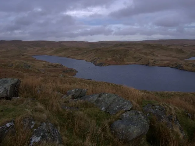 A long, dark upland lake curving through open moorland hills under a cloudy sky, viewed from a rocky rise above its shore.