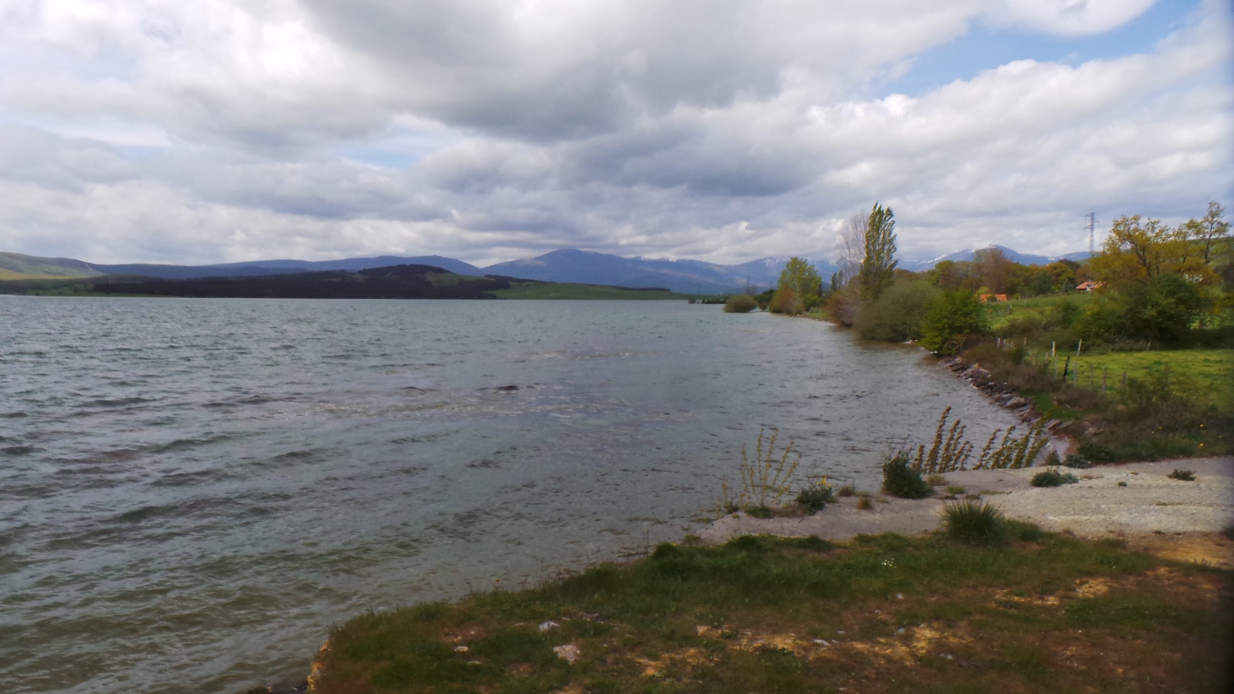 The Ebro river merging into the reservoir in the Orzales area, green hillsides reflected in calm water.