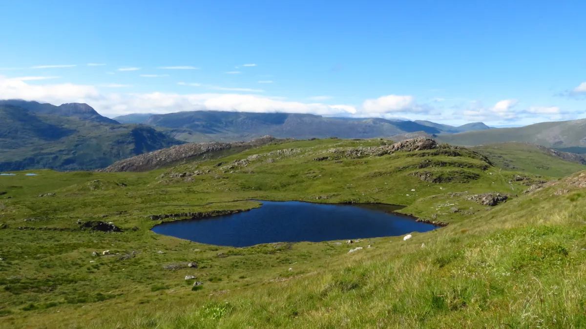 A dark upland lake set in a grassy hollow with rocky outcrops, overlooked by distant Snowdonia mountain ridges under a blue sky.