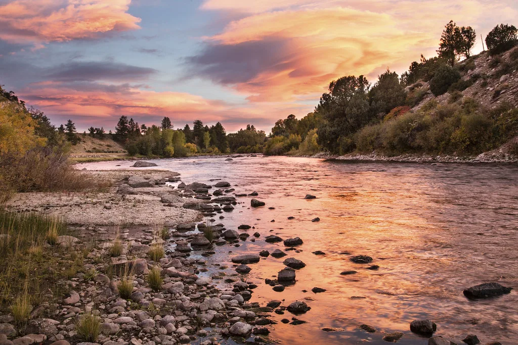 The Arkansas River flowing through a rocky streambed at sunset in Browns Canyon National Monument, Colorado, with pink and orange clouds reflected on the water.