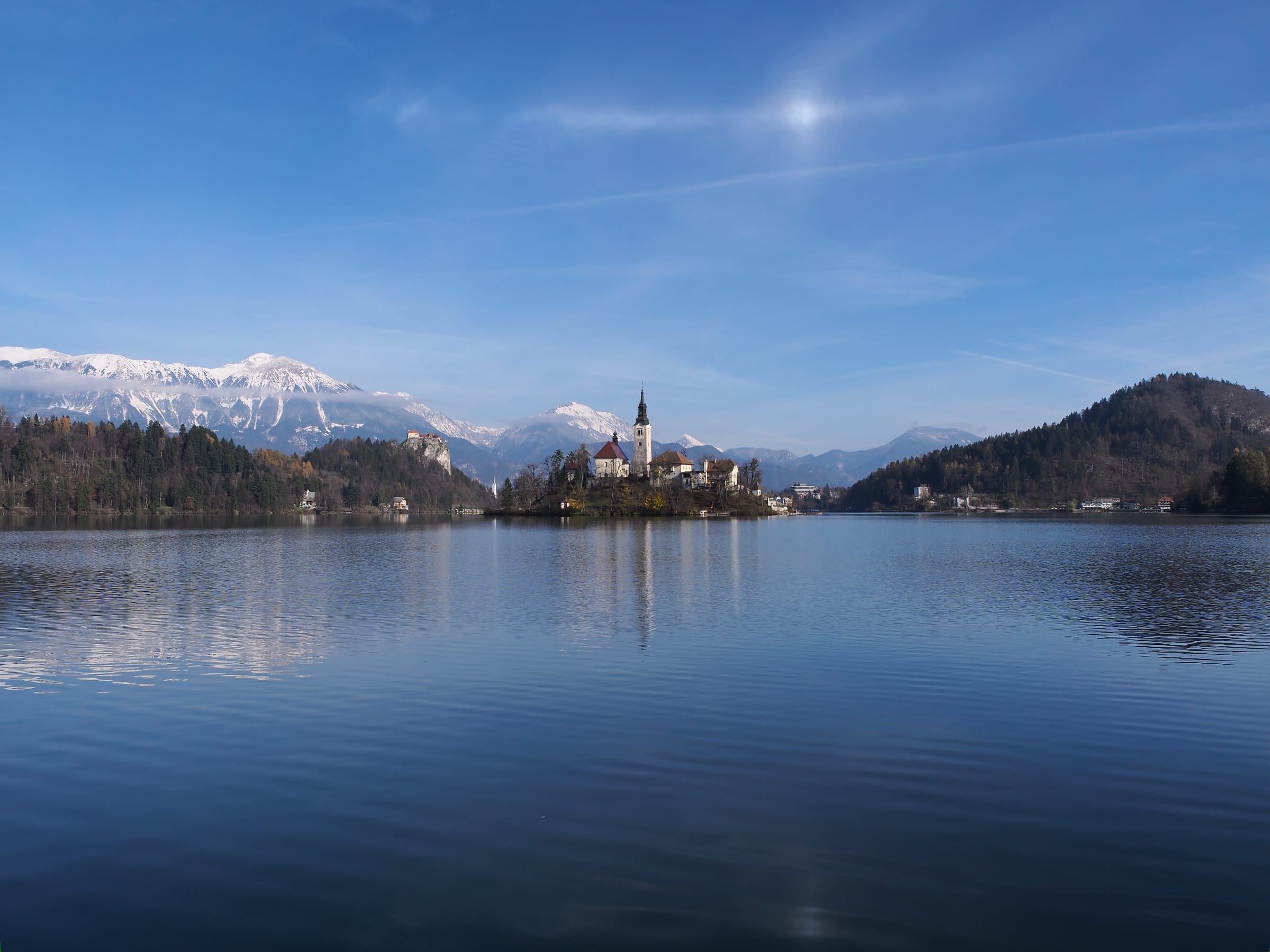 Lake Bled in autumn colours with calm reflective water, the island church mid-lake and forested slopes.