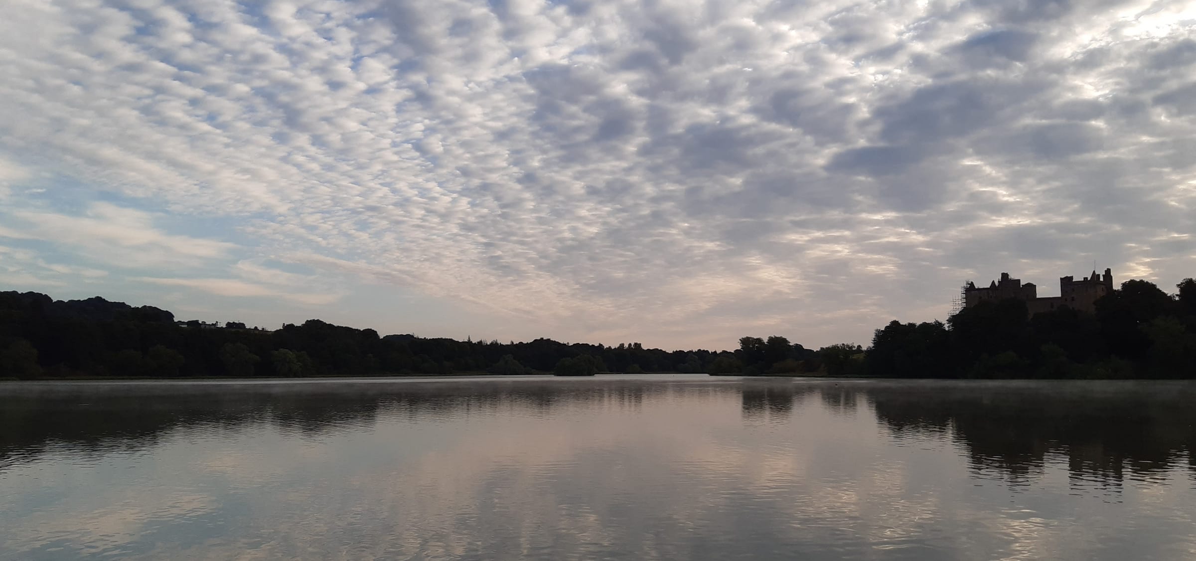 Wide panoramic view of Linlithgow Loch stretching across the frame under a partly cloudy sky, West Lothian.