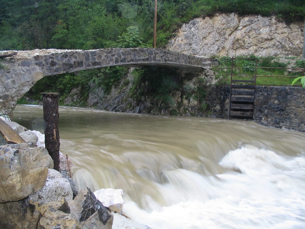 The Trebuščica river in flood at Krtovščah, rushing under a stone arch bridge with limestone gorge walls.