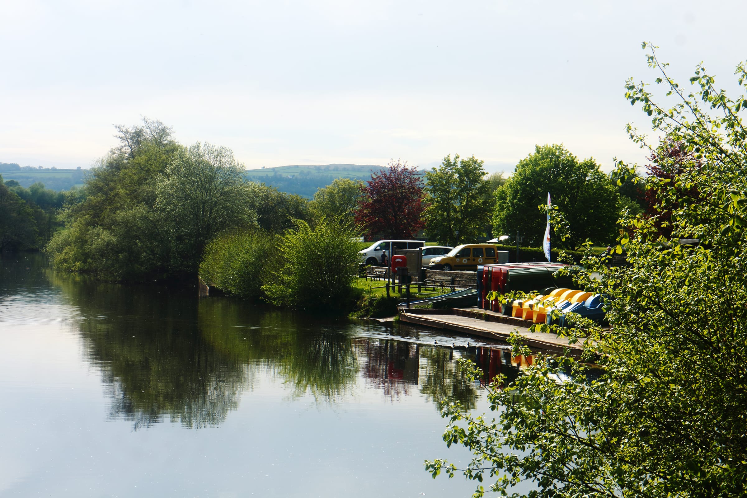 Rowing boats and trees reflected in the clear River Usk at Brecon, with the Brecon Beacons hills beyond.